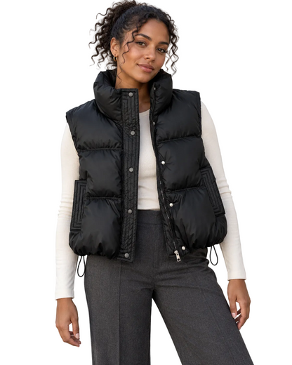 A woman wearing a black puffer vest over a cream long-sleeve top and dark gray trousers, standing outdoors against a textured light beige wall.
