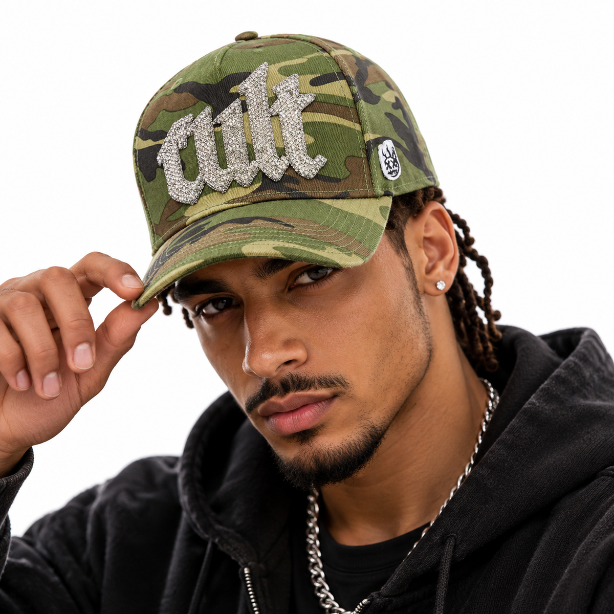 A man wearing a camouflage baseball cap with a large silver rhinestone 'cult' logo on the front, paired with a black hoodie and silver chain necklace against a plain white background.