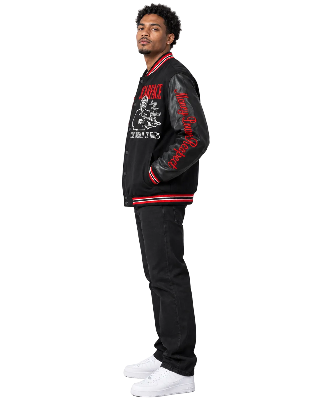 A man stands in profile wearing a black varsity-style jacket with red and white striped ribbing, red script embroidery on the sleeve, and red graphic text on the chest, paired with black jeans and white sneakers against a light gray studio background.