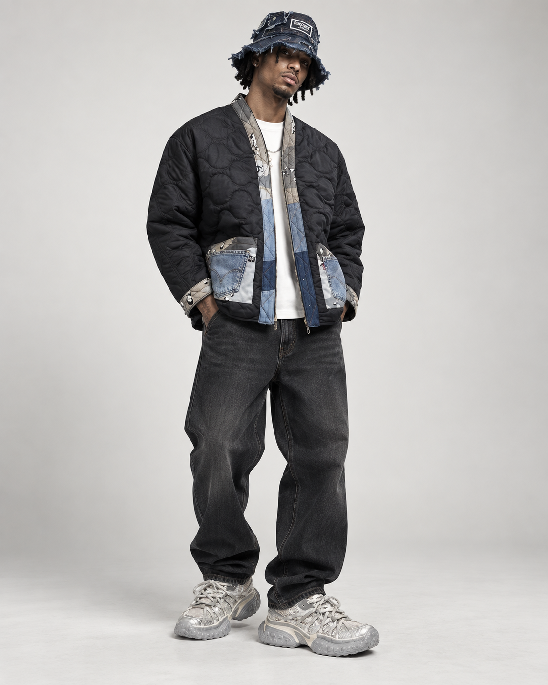 A model stands against a light gray studio background wearing a black quilted bomber jacket with blue denim paneling and silver hardware, a matching bucket hat, dark jeans, and chunky silver sneakers.