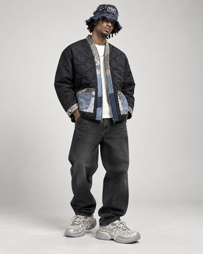 A model stands against a light gray studio background wearing a black quilted bomber jacket with blue denim paneling and silver hardware, a matching bucket hat, dark jeans, and chunky silver sneakers.