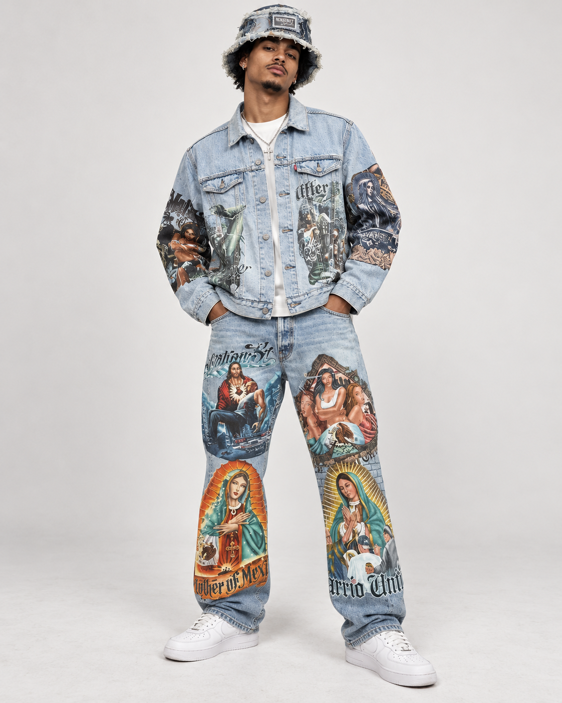 A man modeling a light-wash denim jacket and matching jeans covered in colorful religious and gothic graphic prints, paired with white sneakers and a bucket hat, against a plain light gray studio background.