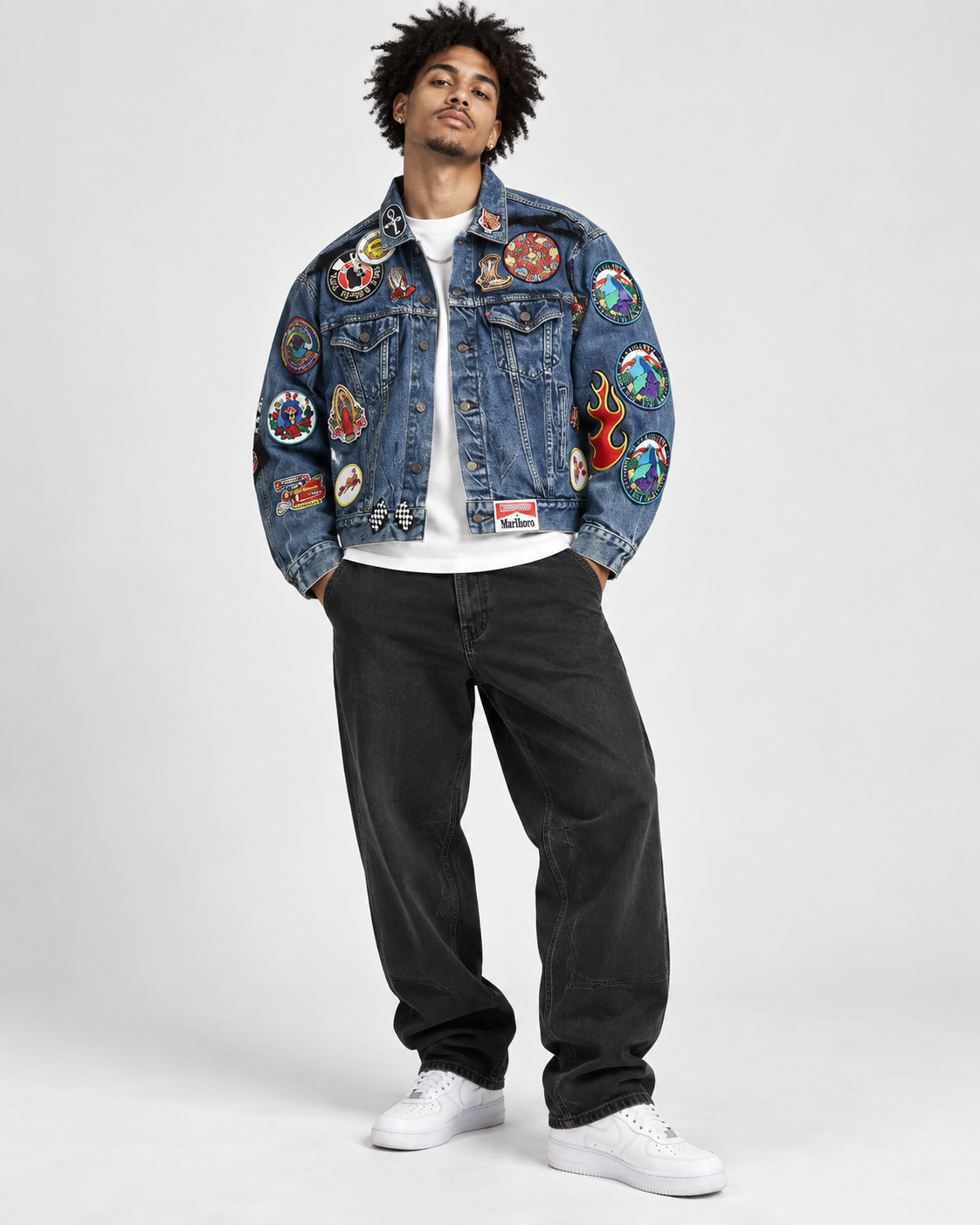 A man modeling a heavily decorated blue denim jacket with colorful embroidered patches, worn over a white t-shirt and black jeans, on a light gray studio background.
