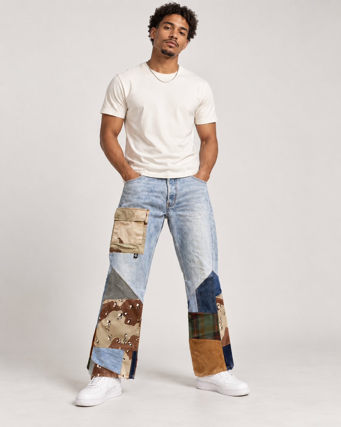 A man modeling a pair of light blue patchwork jeans with various colored and patterned fabric panels, paired with a white t-shirt and white sneakers, against a light gray studio background.
