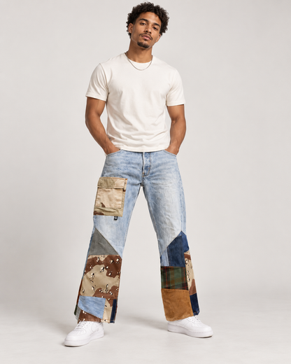 A man modeling a pair of light blue patchwork jeans with various colored and patterned fabric panels, paired with a white t-shirt and white sneakers, against a light gray studio background.