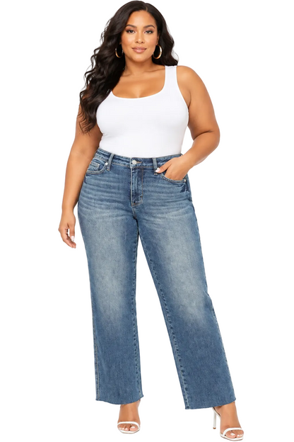 A woman modeling a pair of high-waisted, wide-leg blue jeans with a white tank top and white high-heeled sandals, standing against a plain white background.