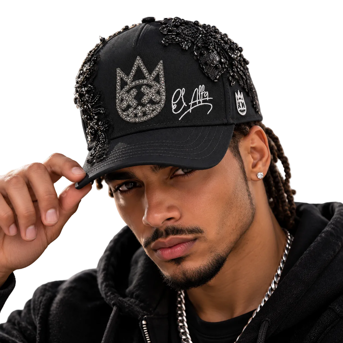 A man wearing a black baseball cap adorned with rhinestone crown and script embroidery, embellished with black beaded fringe on the sides, in a close-up portrait.