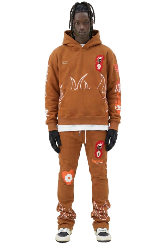 A model wearing a matching brown hoodie and sweatpants set with white flame graphics, red and white patches, a white undershirt, black gloves, and white sneakers, set against a plain white background.