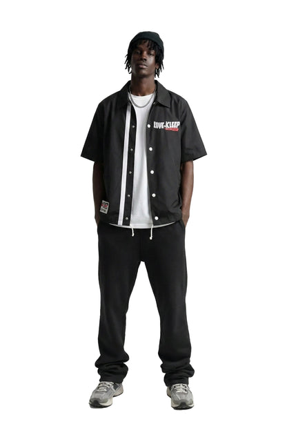 A model stands against a solid white background wearing a black short-sleeve button-up jacket with white stripes and a white 'LOVE-KEEP' logo on the chest, layered over a white t-shirt, black sweatpants, and gray sneakers.