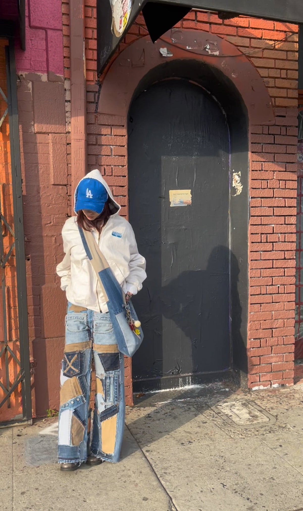 A person stands on a sidewalk in front of a red brick building with a dark arched door, wearing a white hooded jacket, light blue patchwork wide-leg jeans, and a blue baseball cap.