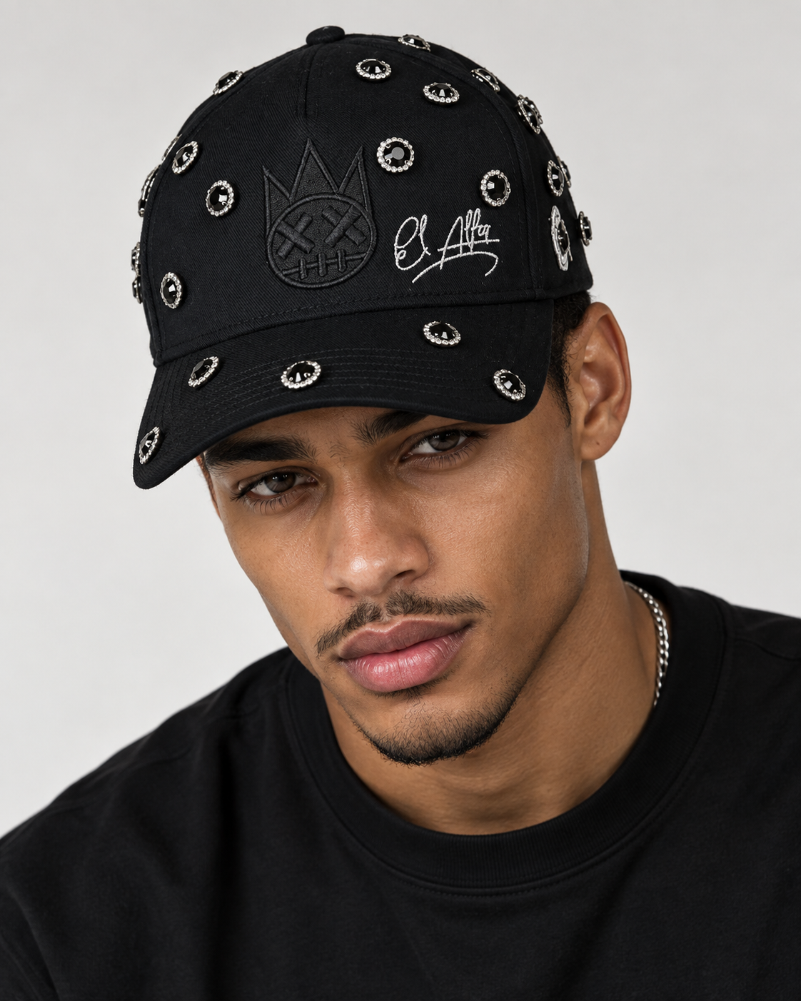 A close-up of a man wearing a black baseball cap adorned with silver eye studs and embroidered details, including a crown logo and script.