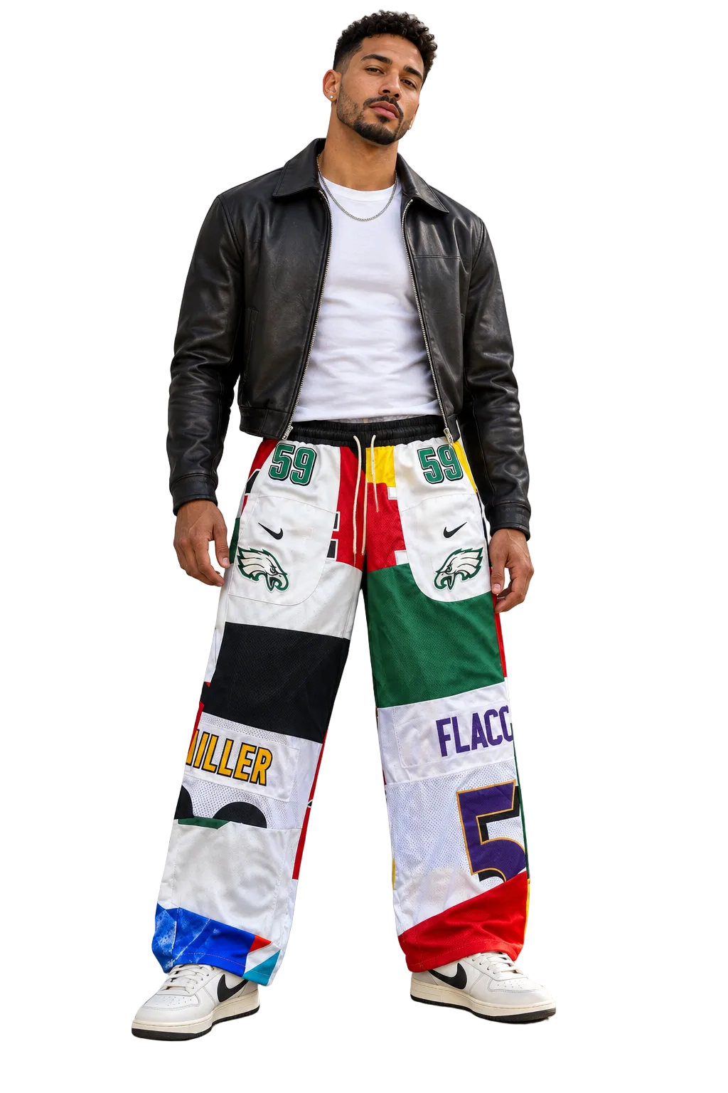A man wearing a black leather jacket, white t-shirt, and a pair of highly colorful, patchwork-style wide-leg pants with sports team graphics, standing against a concrete wall.