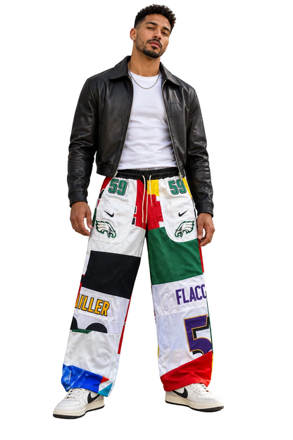A man wearing a black leather jacket, white t-shirt, and a pair of highly colorful, patchwork-style wide-leg pants with sports team graphics, standing against a concrete wall.