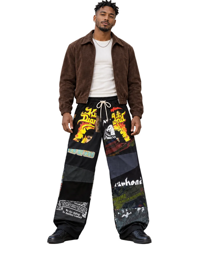 A man stands outdoors against a light stone building wall wearing baggy black patchwork pants with colorful graphic prints, a brown suede jacket, a white t-shirt, and black shoes.