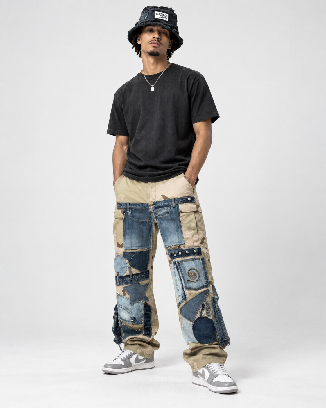 A man modeling a pair of wide-leg, patchwork-style cargo pants made from various blue and beige denim jeans, styled with a black t-shirt, a black bucket hat, and gray sneakers against a light gray studio background.