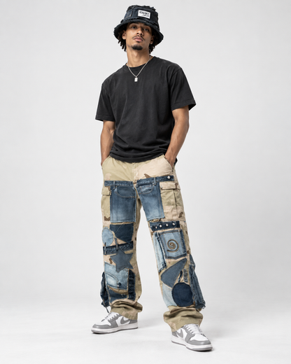 A man modeling a pair of wide-leg, patchwork-style cargo pants made from various blue and beige denim jeans, styled with a black t-shirt, a black bucket hat, and gray sneakers against a light gray studio background.