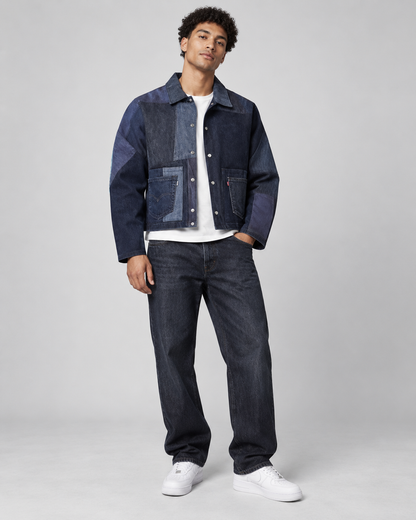 A man modeling a dark blue patchwork denim jacket with a white t-shirt, dark blue jeans, and white sneakers, standing against a light gray studio background.