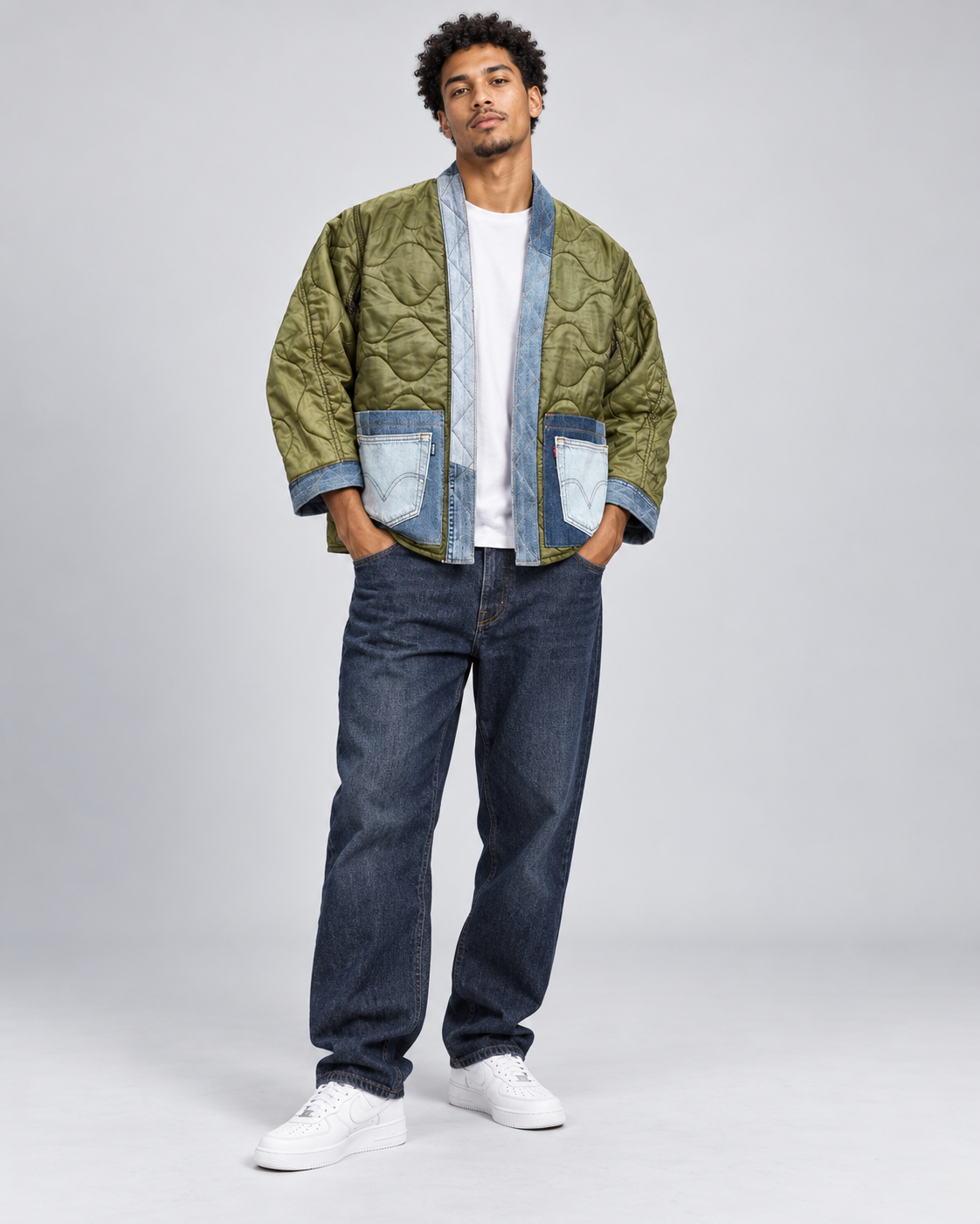 A man modeling a unique quilted bomber jacket made from olive green and light blue denim panels, worn over a white t-shirt and dark blue jeans, on a light gray studio background.