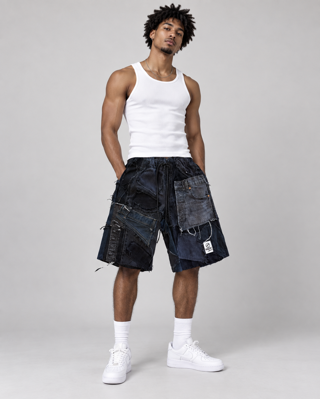 A male model wearing dark blue patchwork denim shorts, a white ribbed tank top, white socks, and white sneakers, standing against a light gray studio background.