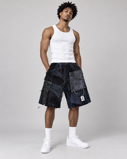 A male model wearing dark blue patchwork denim shorts, a white ribbed tank top, white socks, and white sneakers, standing against a light gray studio background.