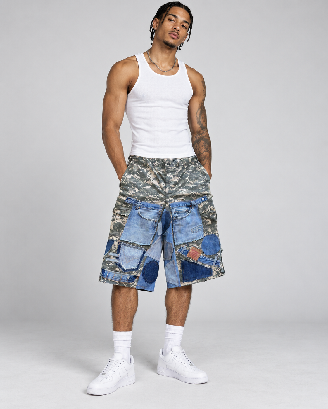 A man modeling a pair of cargo shorts featuring a blue denim patchwork print over a camouflage base, styled with a white tank top and white sneakers against a light gray studio background.