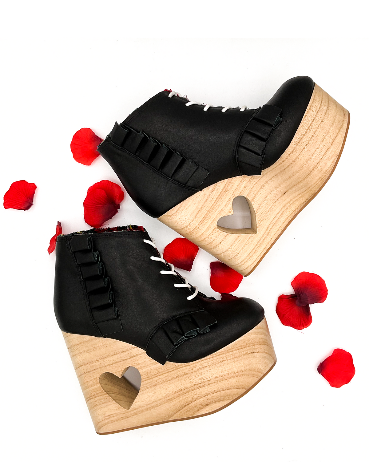 A pair of black platform ankle boots with ruffle trim, white laces, and a heart-shaped cutout on the wooden wedge, styled with scattered red rose petals on a white background.
