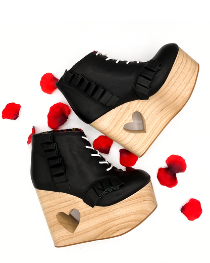 A pair of black platform ankle boots with ruffle trim, white laces, and a heart-shaped cutout on the wooden wedge, styled with scattered red rose petals on a white background.