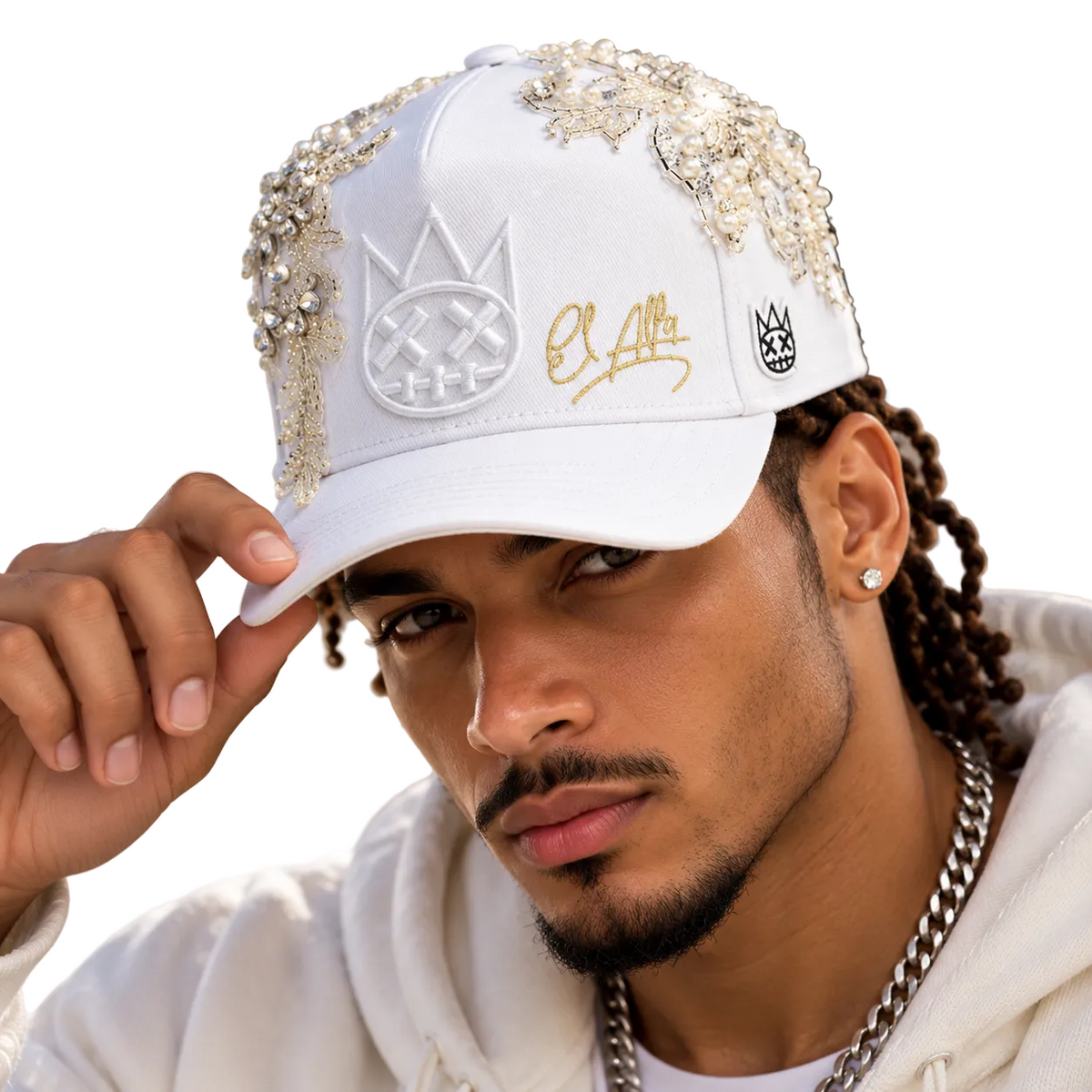 A man wearing a white baseball cap embellished with rhinestones, gold script embroidery, and a black-and-white crown logo, posed outdoors against a blurred architectural background.