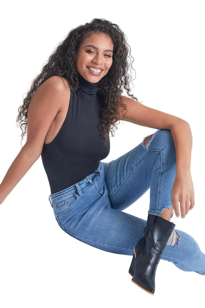 A woman sitting on a light gray studio floor wearing a sleeveless black mock-neck top, distressed blue jeans, and black ankle boots.
