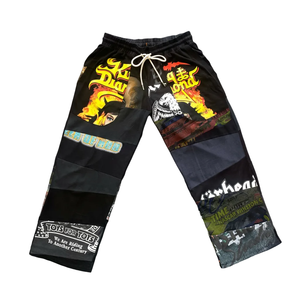 A pair of black patchwork cargo-style pants with a white drawstring, featuring various graphic prints, text, and collage elements, laid flat on a light gray concrete surface.