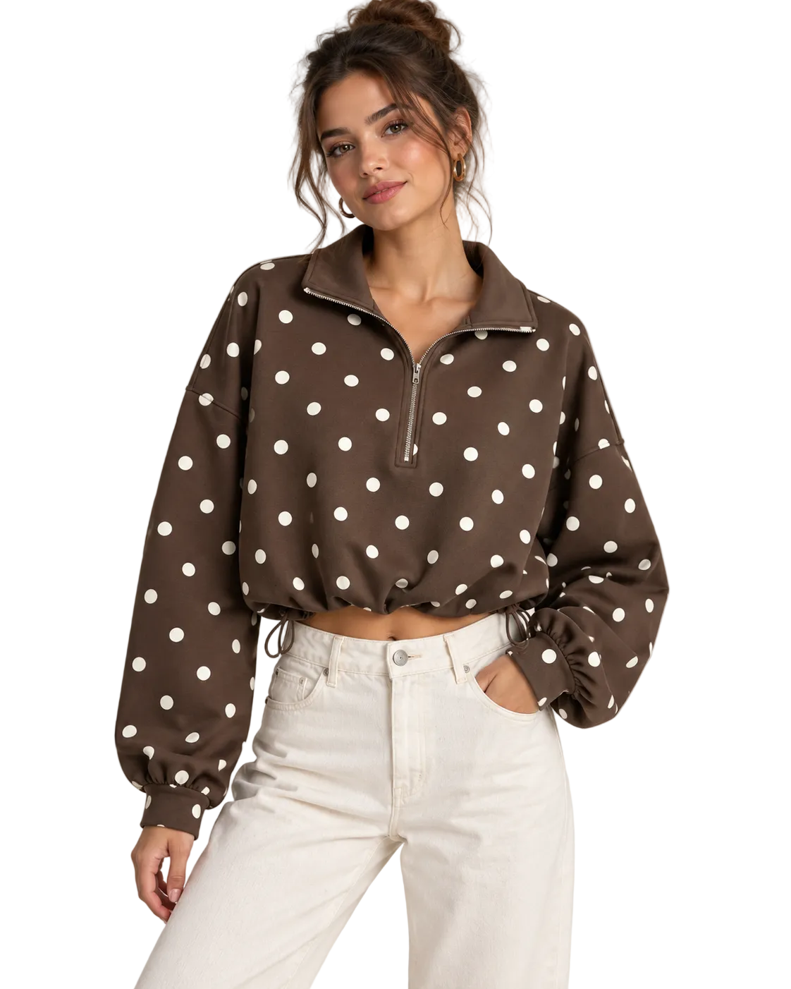 A woman wearing a brown long-sleeve quarter-zip pullover with white polka dots, paired with white jeans, against a plain beige studio background.