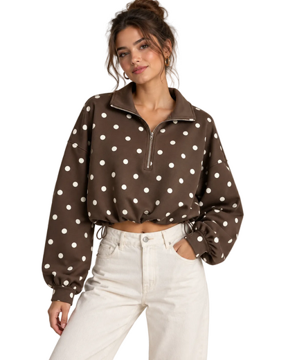 A woman wearing a brown long-sleeve quarter-zip pullover with white polka dots, paired with white jeans, against a plain beige studio background.