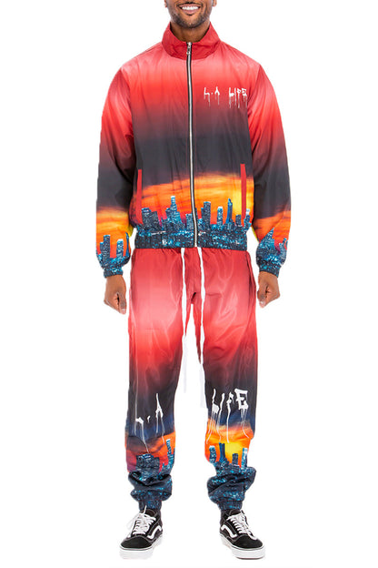 A man modeling a matching zip-up jacket and jogger pants set with a vibrant red, orange, and black gradient cityscape print, shown against a plain white background.