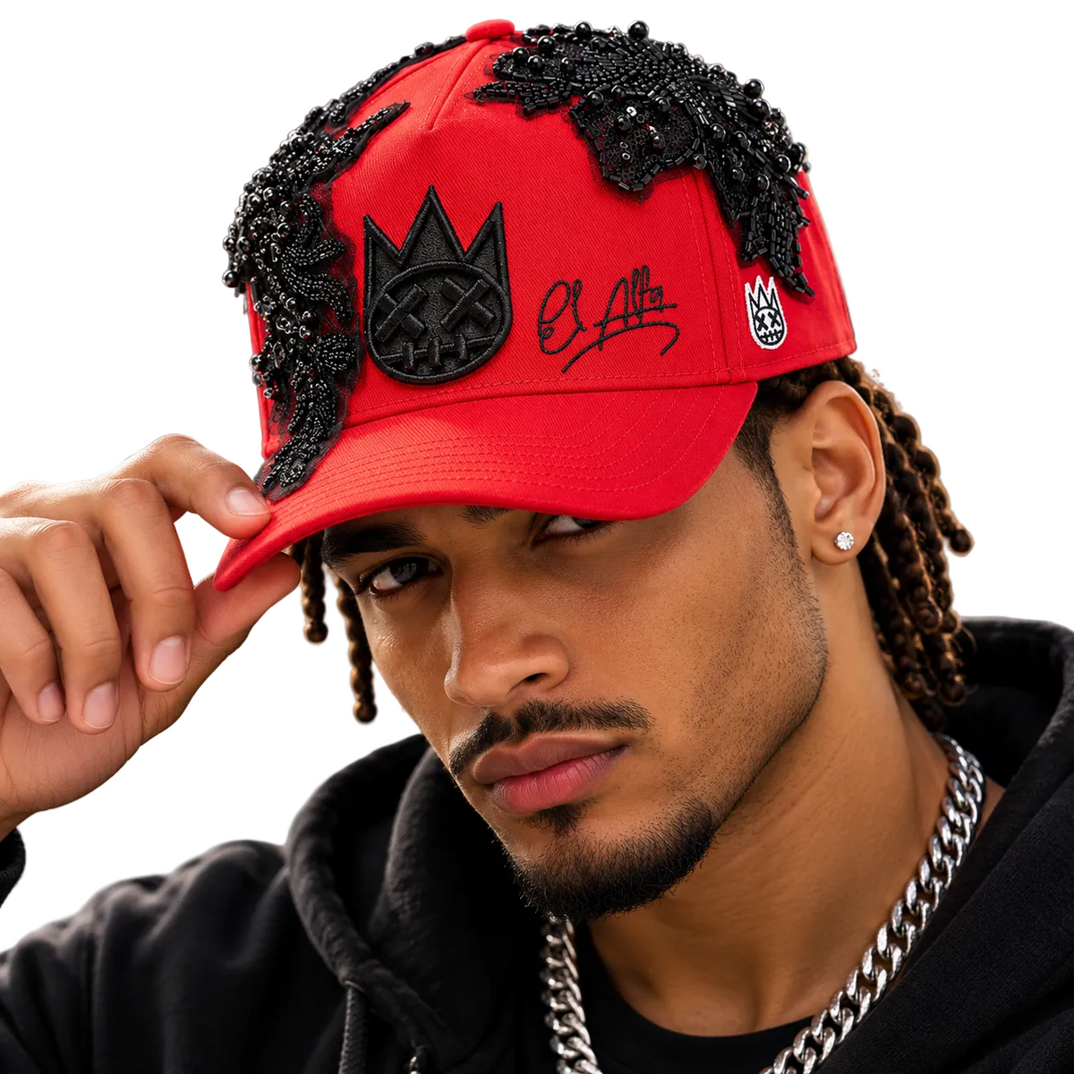 A close-up lifestyle shot of a man wearing a bright red baseball cap adorned with black beaded lace details, a black crown patch with a skull design, and a black script logo on the front. He is wearing a black hoodie and a silver chain necklace against a blurred city background.
