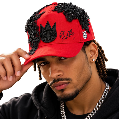 A close-up lifestyle shot of a man wearing a bright red baseball cap adorned with black beaded lace details, a black crown patch with a skull design, and a black script logo on the front. He is wearing a black hoodie and a silver chain necklace against a blurred city background.