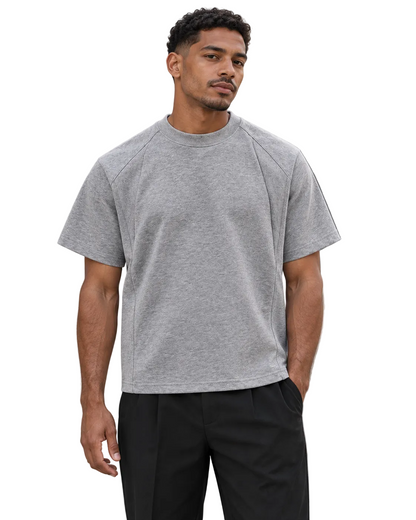 A man wearing a light gray short-sleeve crewneck t-shirt and black pants, standing outdoors against a concrete wall.