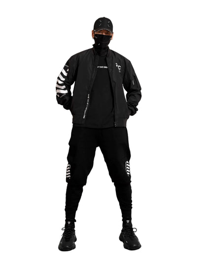 A model wearing a black streetwear outfit consisting of a bomber jacket with white sleeve stripes, a black t-shirt, black cargo pants with white thigh stripes, a black cap, and a black face mask, standing against a gray studio background.