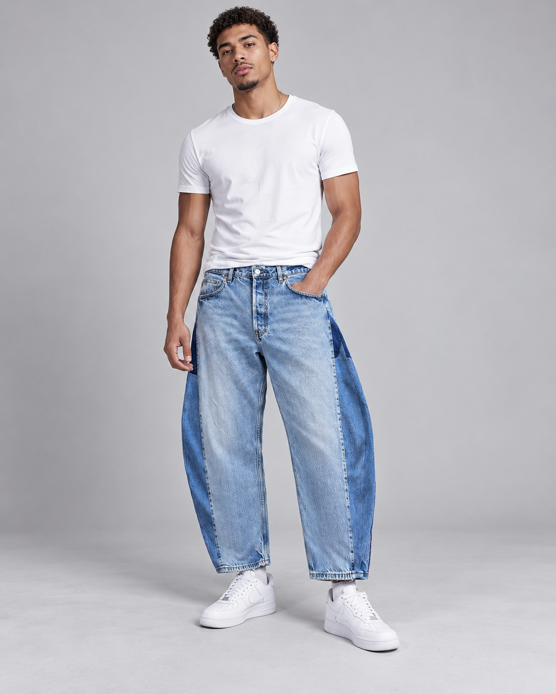 A man modeling a pair of wide-leg light blue denim jeans with dark blue contrast side panels, styled with a white t-shirt and white sneakers against a light gray studio background.