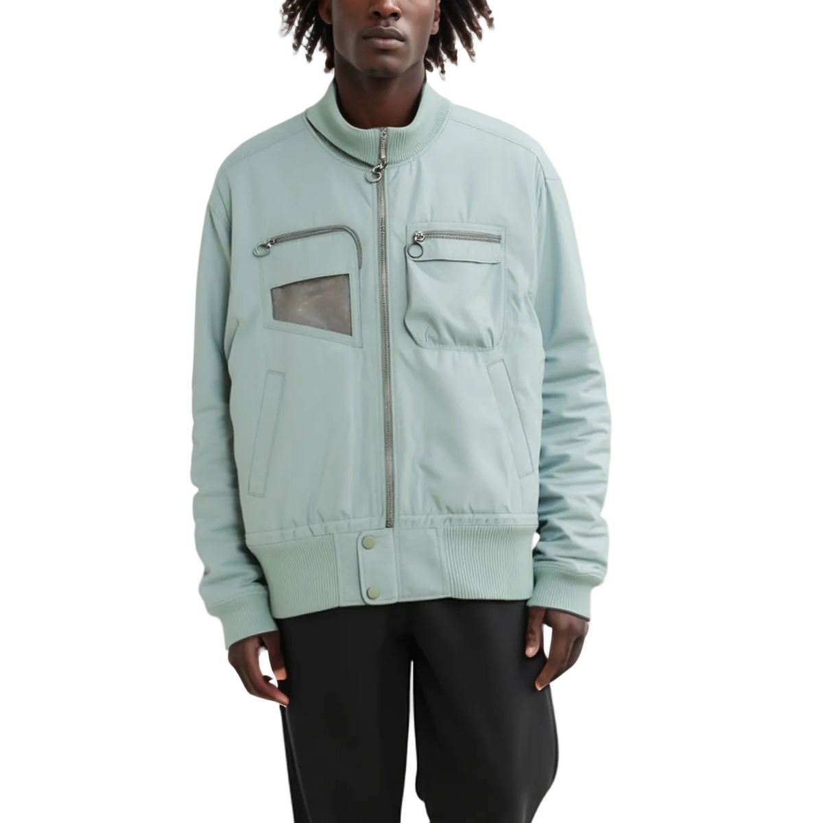A model wearing a light sage green bomber jacket with silver zippers and a transparent window on the chest pocket, paired with black pants, against a plain white background.