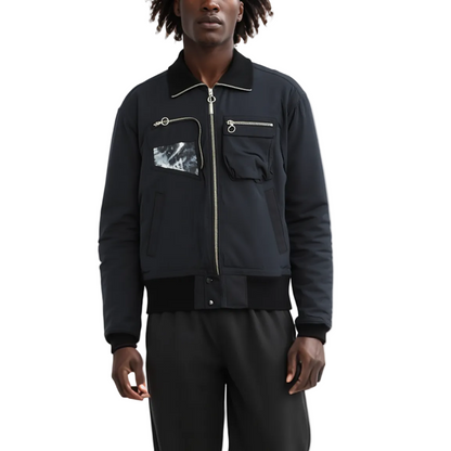 A man wearing a black bomber jacket with a silver zipper, a black-and-white marble patterned chest pocket, and black pants, photographed against a plain white studio background.