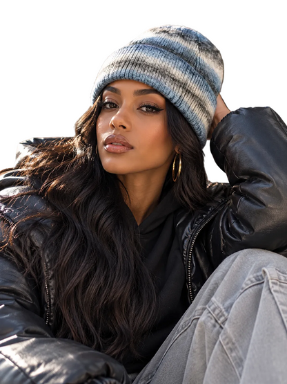 A woman wearing a blue and gray striped knit beanie, a black puffer jacket, and light gray jeans, posing outdoors against a stone building background.