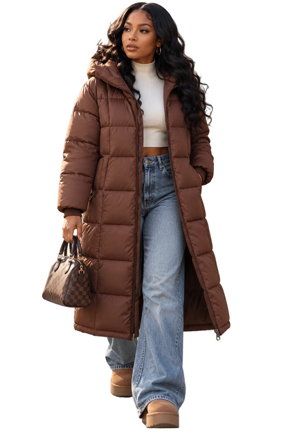 A woman walking outdoors wearing a long brown puffer coat over a white cropped top and light blue wide-leg jeans, holding a brown patterned handbag.