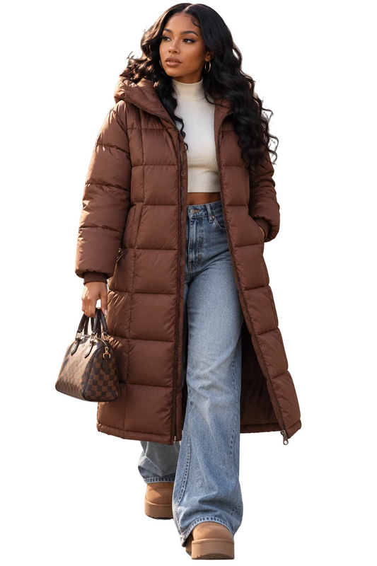 A woman walking outdoors wearing a long brown puffer coat over a white cropped top and light blue wide-leg jeans, holding a brown patterned handbag.