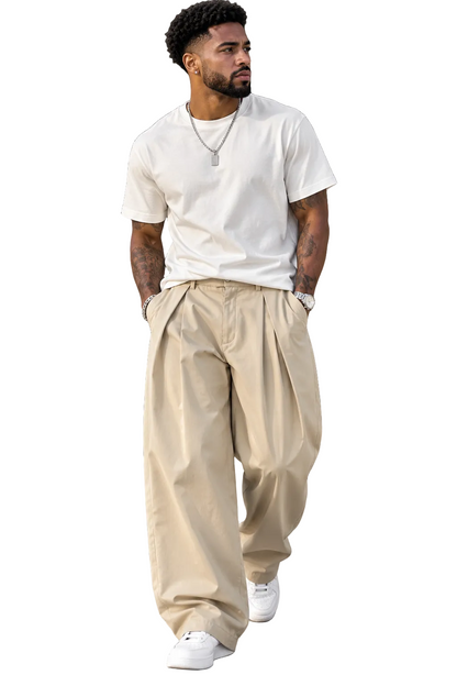 A man walking outdoors wearing a white crew-neck t-shirt, loose beige pleated trousers, and white sneakers.
