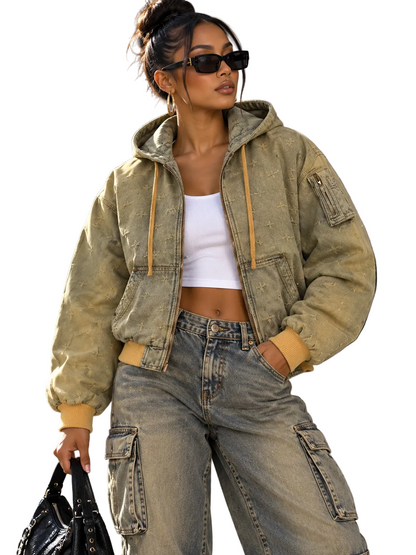 A woman wearing a faded olive green hooded bomber jacket with tan drawstrings and cuffs, styled with light-wash cargo jeans, a white crop top, and black sunglasses, holding a black handbag in an outdoor urban setting.