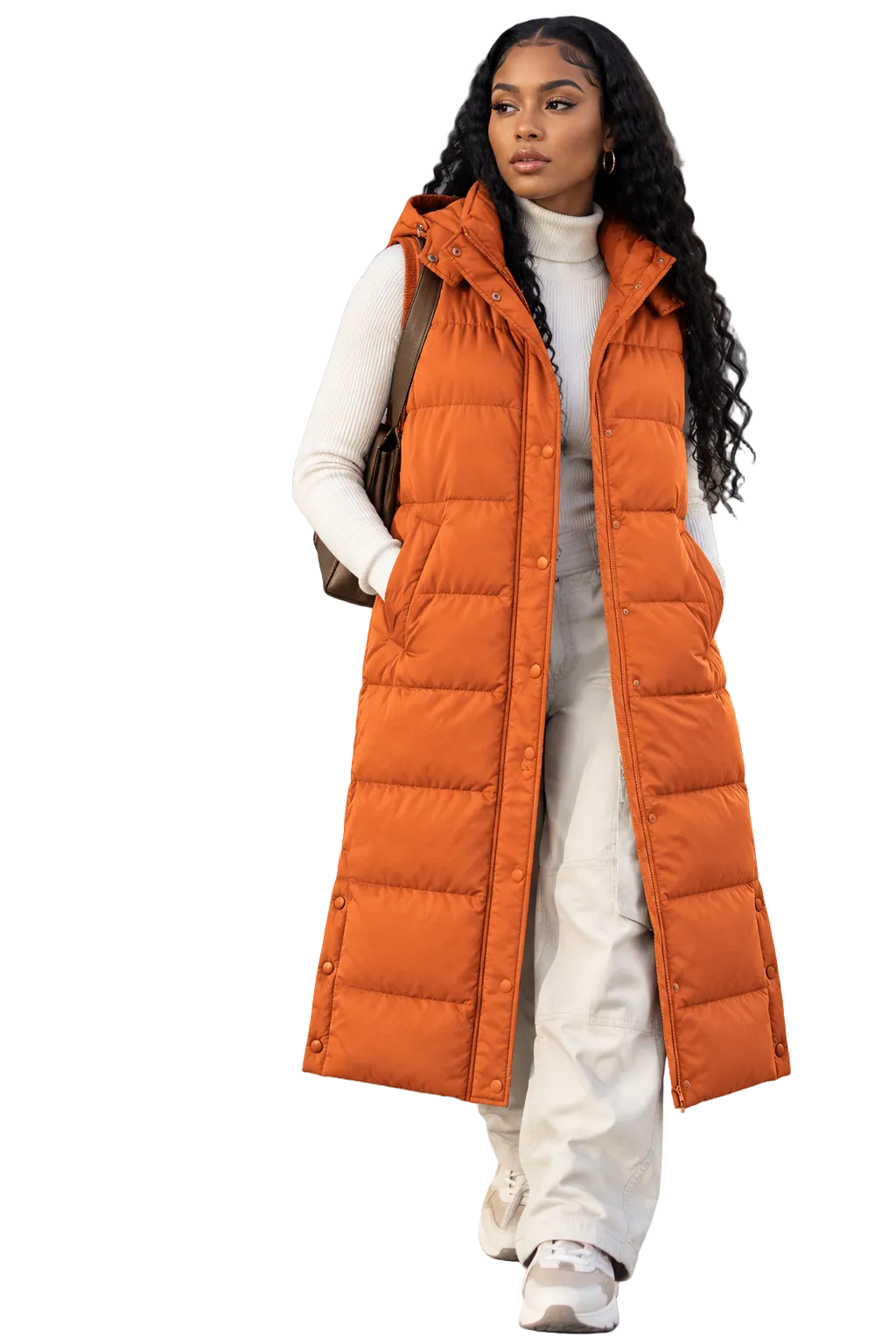 A woman modeling a long orange puffer vest over a white turtleneck and white pants, standing outdoors against a gray concrete wall. The brand name KRIFFA is printed in white at the top left.
