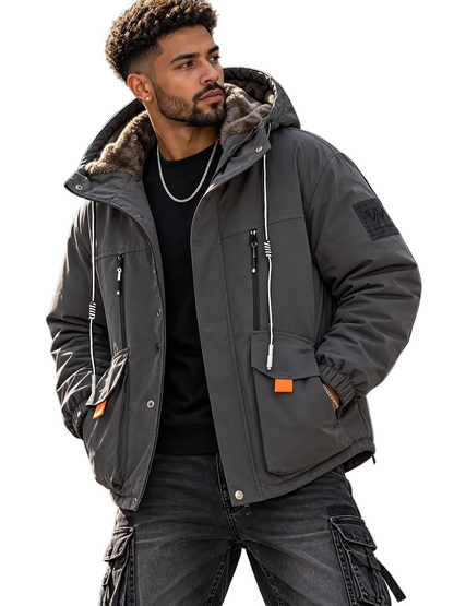 A man wearing a dark gray hooded puffer jacket with a brown faux-fur lined hood, black cargo pants, and a black t-shirt, standing on outdoor concrete steps.