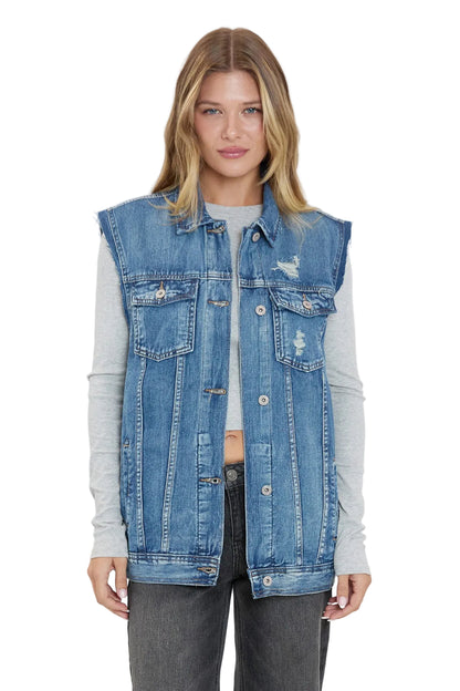 A woman modeling a sleeveless light blue denim vest over a light gray long-sleeve shirt and dark gray jeans, photographed against a plain light gray studio background.