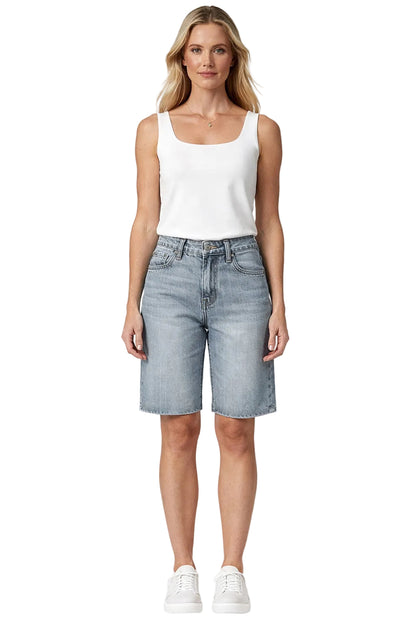 A woman modeling a white sleeveless tank top and light-wash denim shorts against a plain light gray studio background.