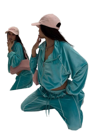 A woman kneels on a white studio floor wearing a turquoise velvet matching lounge set with white piping, a pink baseball cap, and a pink shoulder bag, with her reflection visible in a large mirror.